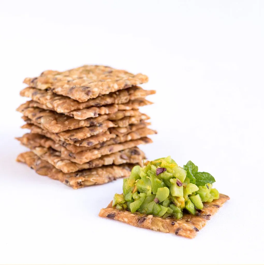 Mette is Baking crackers stacked on top of one another in the background, with a Mette is Baking Nordic cracker in the foreground topped with edamame bean and avocado dip