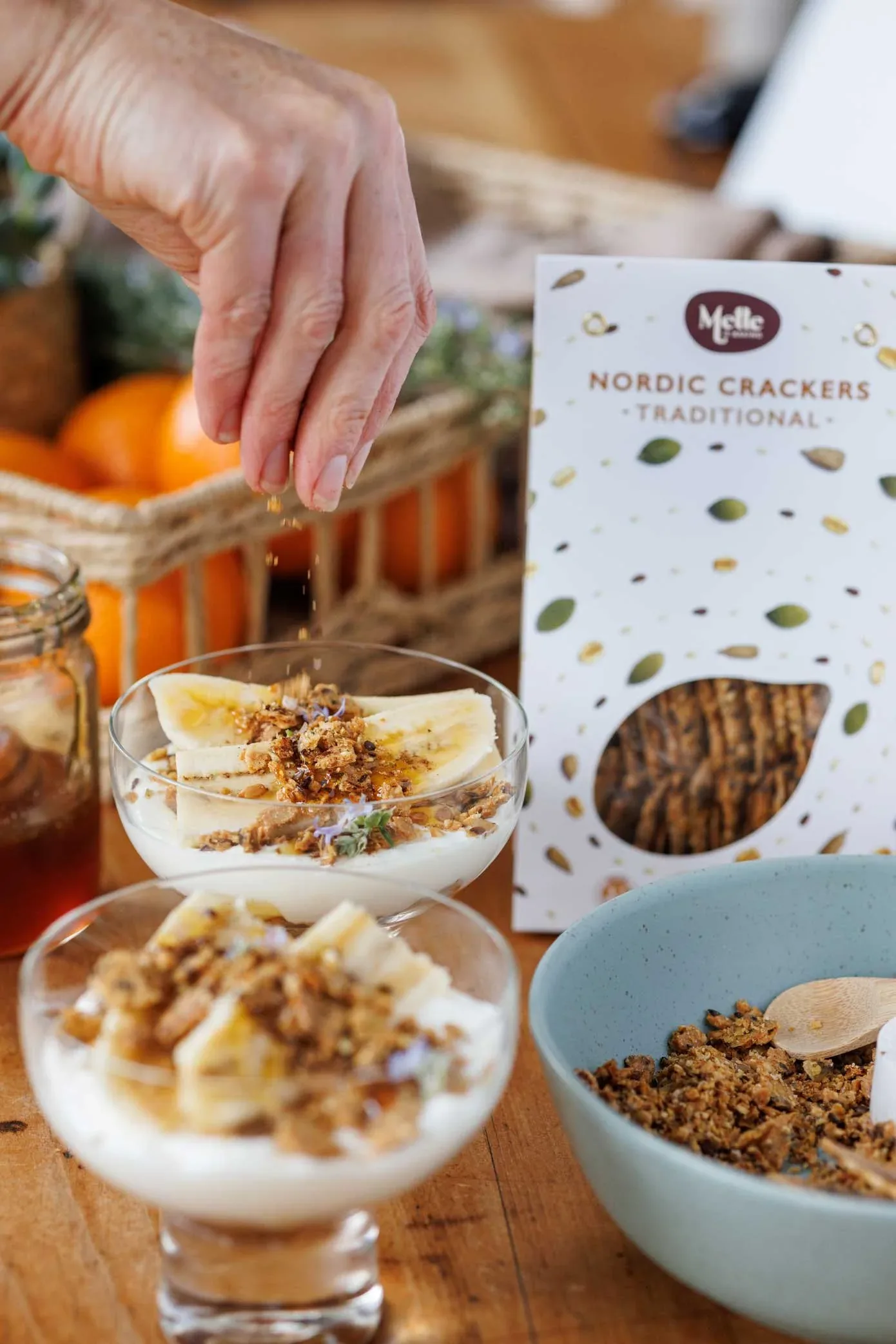 Two glass bowls filled with banana slices and Greek yoghurt, with Mette is Baking cracker crumbs being sprinkled on top