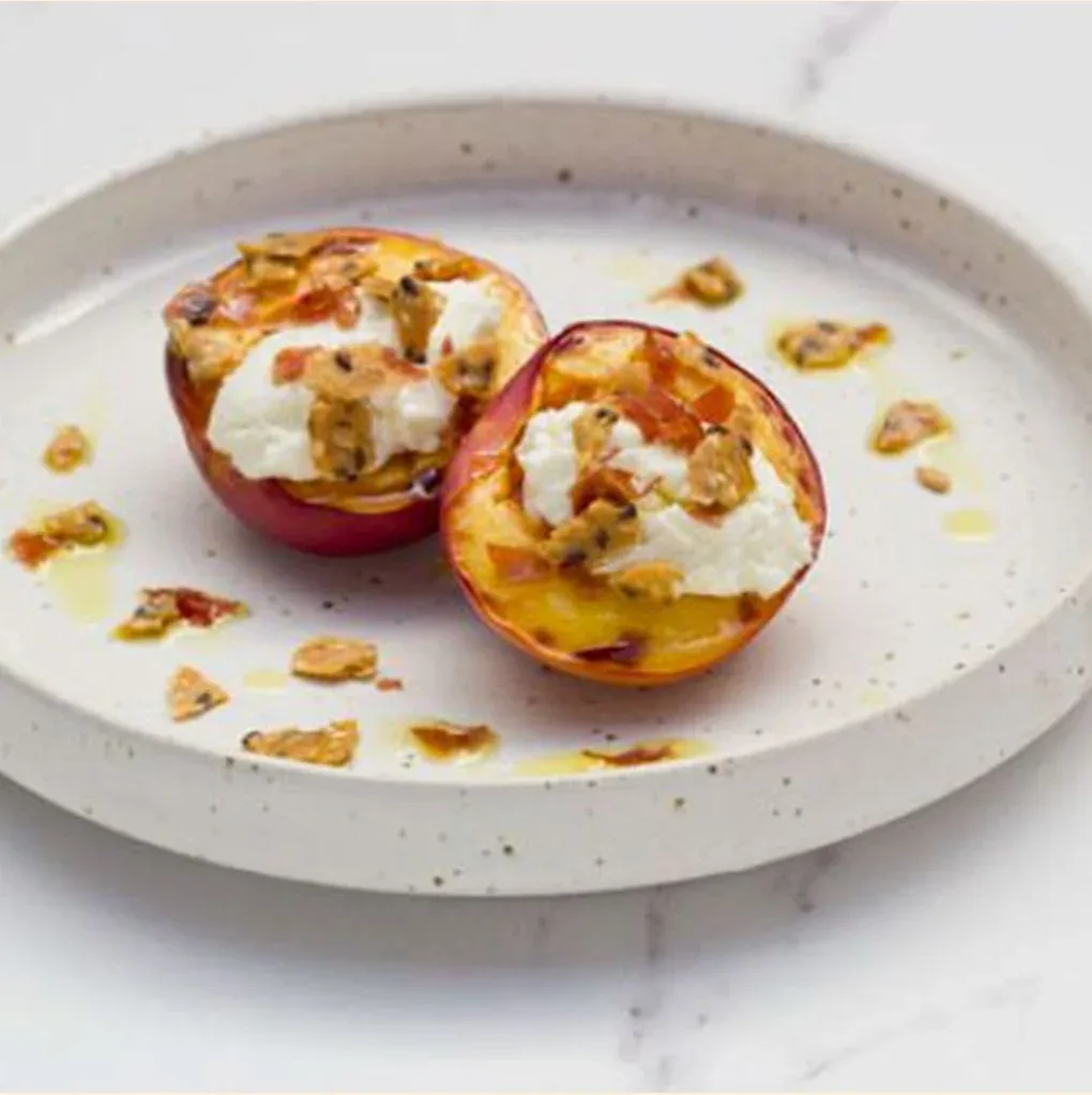 Two halves of a nectarine on a serving plate, grilled and topped with goat chevre and Mette is Baking Nordic cracker crumbs