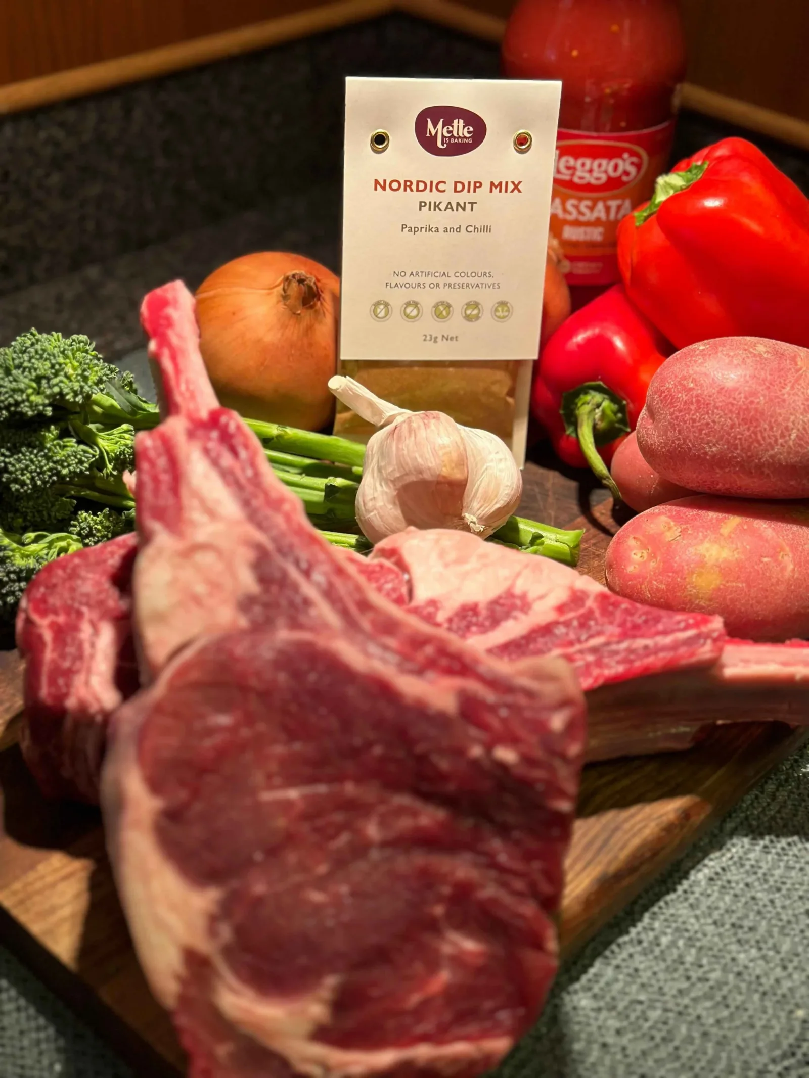 Two raw ribeye steaks with all the ingredients needed for the slow cooked ribeye recipe, including onions, capsicum, Mette is Baking Pikant Dip Mix, garlic, potatoes and broccolini