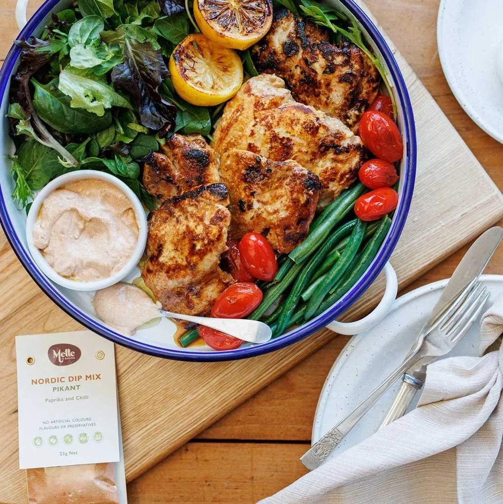 A flat lay image of a large white serving bowl on a wooden table, filled with lettuce leaves, a lemon cut in half, marinated chicken in pikant spices, steamed green beans and roasted cherry tomatoes
