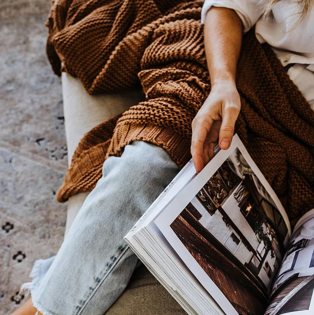 A lady sitting on a couch with a brown knitted blanket over her legs, reading a book feeling warm and cosy in winter
