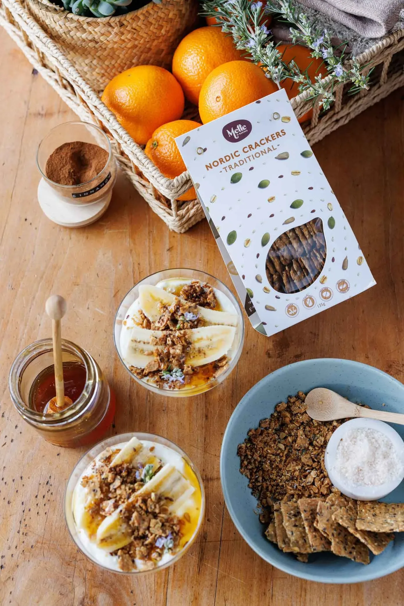 A wooden dining table with a jar of honey, cinnamon, oranges, a pack of Mette is Baking crackers, two bowls filled with Greek yoghurt and banana and a blue bowl filled with Mette is Baking crackers and cracker crumbs