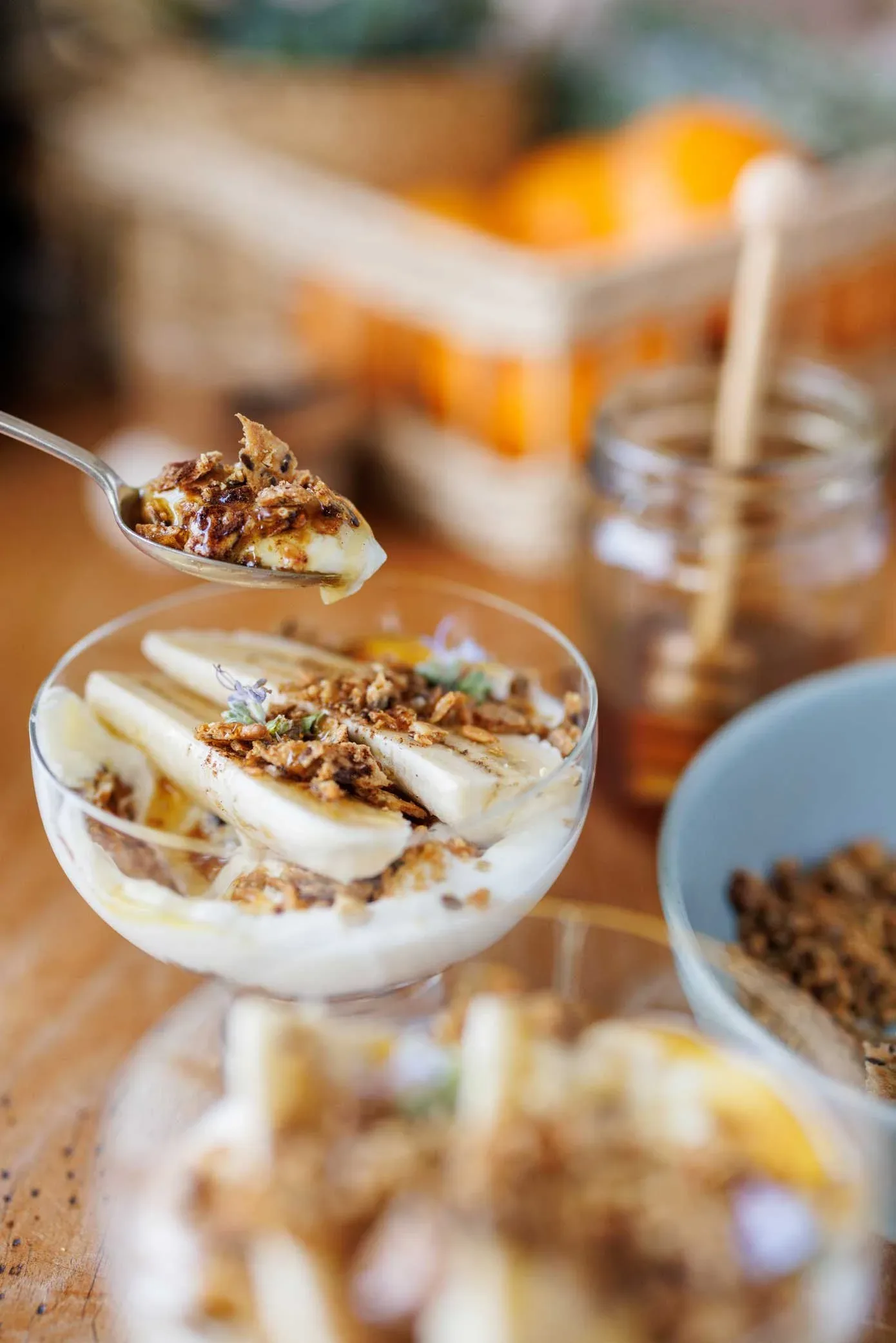 Two glass bowls filled with Greek yoghurt, banana and Mette is Baking cracker crumbs topped with honey and a silver spoon showcasing the ingredients