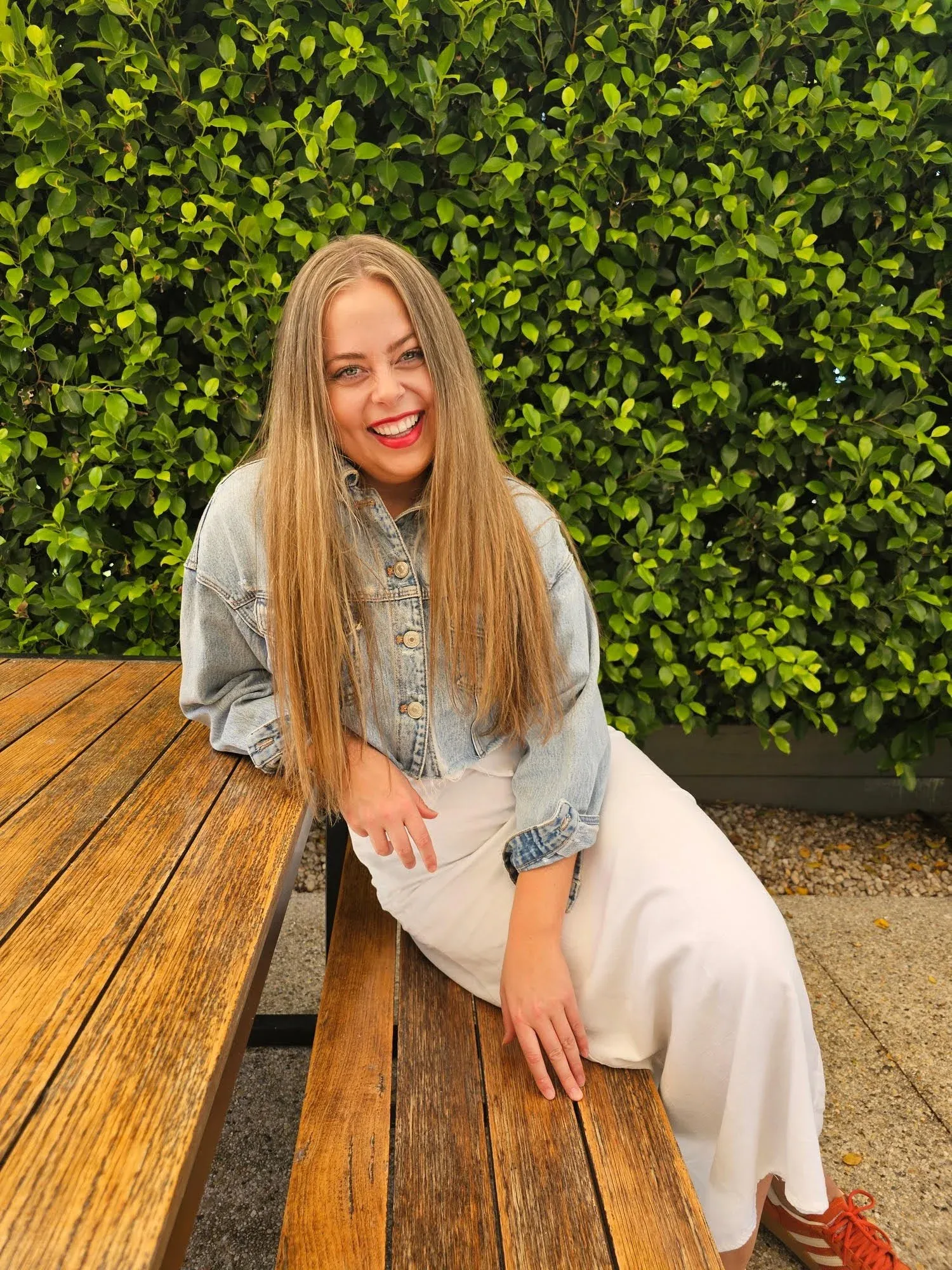 Alex Crisp sitting on a chair and leaning against a table outside, pictured in front of a large green plant