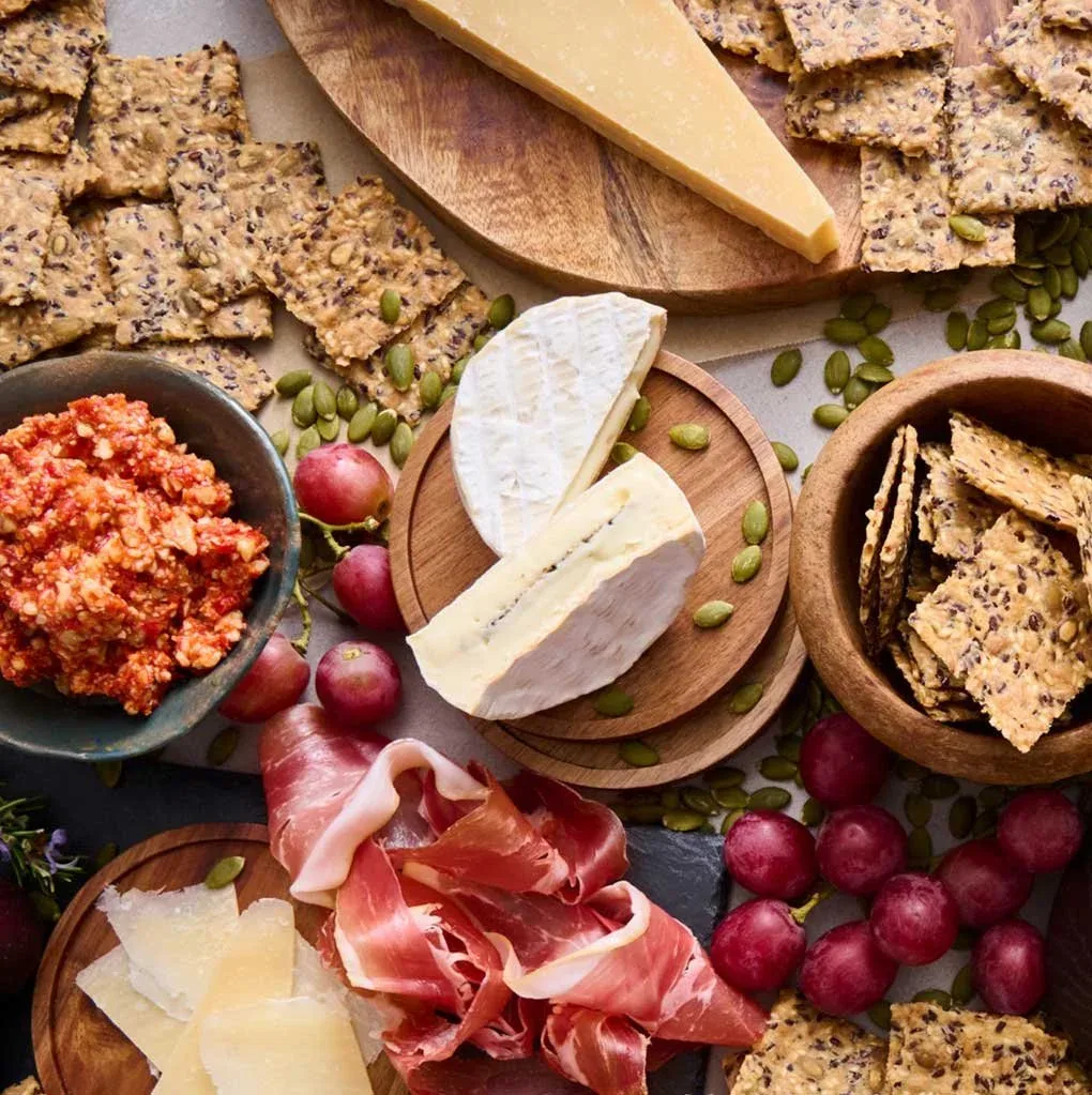 A flat lay image of a grazing board filled with different types of cheeses, dips, cold cut meats, grapes, pumpkin seeds and Mette is Baking crackers